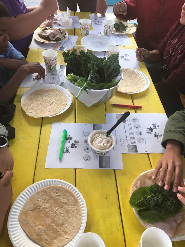 A resident-led lesson on healthy recipes and safe food handling at REACH site Alliance Academy – students making wraps and pineapple salsa using kale, lettuce, tomatoes, and herbs from the school garden
